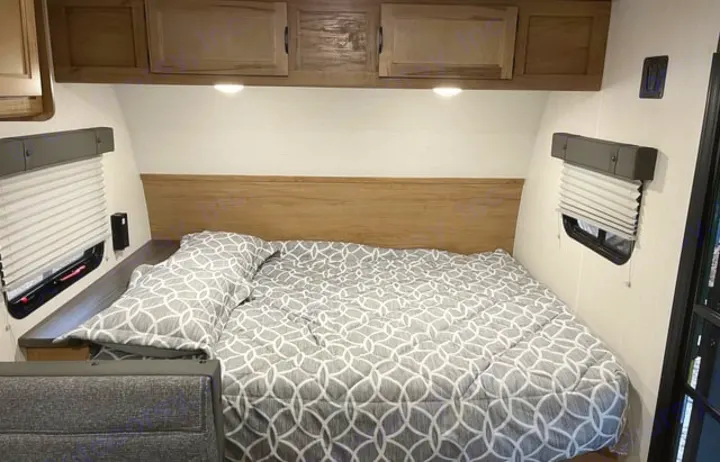 Experience comfort in this RV rental bedroom, featuring a cozy bed, wooden headboard, overhead cabinets, and two windows.. Forest River Cherokee Wolf Pup 2024