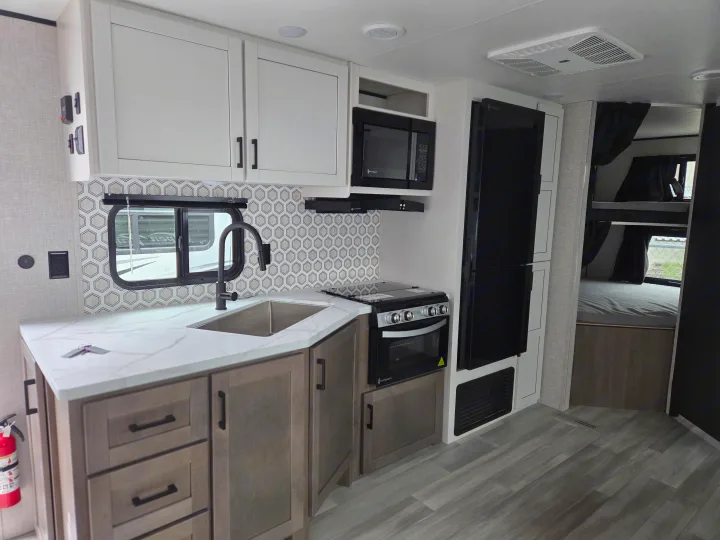 Modern RV rental kitchen with white cabinets, full appliances (stove, microwave, fridge), sink, and bunk beds visible in the background.. Open Range Conventional 26BHS Travel Trailer 2025