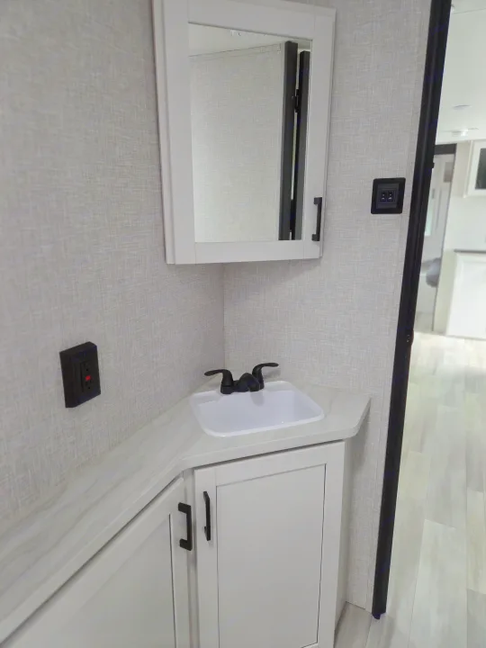 Corner vanity in an RV rental bathroom, featuring a white sink, black faucet, white mirrored medicine cabinet, and textured gray walls.. Open Range Conventional 26BHS Travel Trailer 2025