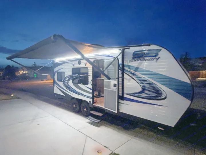 This bright RV rental travel trailer, with its awning extended and lights on at dusk, stands ready on a driveway for new camping adventures.. Pacific Coachworks Sandsport 2017