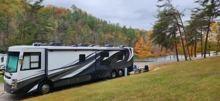 A large RV rental is parked by a tranquil lake, nestled amidst hills adorned with vivid autumn foliage. A picnic area is nearby.. Forest River Berkshire XLT 2024