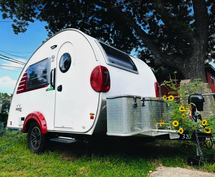 White teardrop RV rental with red accents, parked under a large tree with sunflowers. Perfect for outdoor adventures!. nuCamp T@B 2022