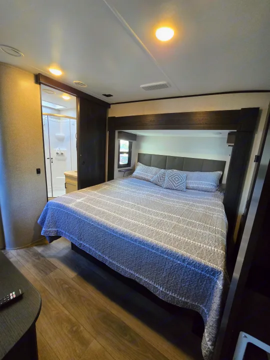 Comfortable RV rental bedroom with a patterned bedspread, dark wood accents, wood-look flooring, and a peek into the bright bathroom.. Highland Ridge RV Mesa Ridge 2021