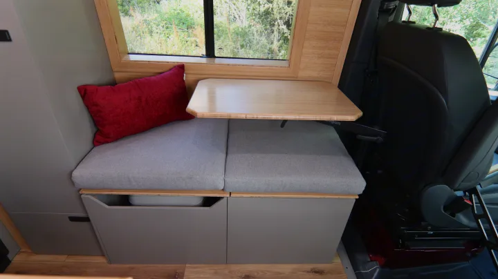 RV rental's inviting camper van interior: bench with red pillow, bamboo table, storage, and a scenic window view.. Mercedes-Benz Sprinter 2025