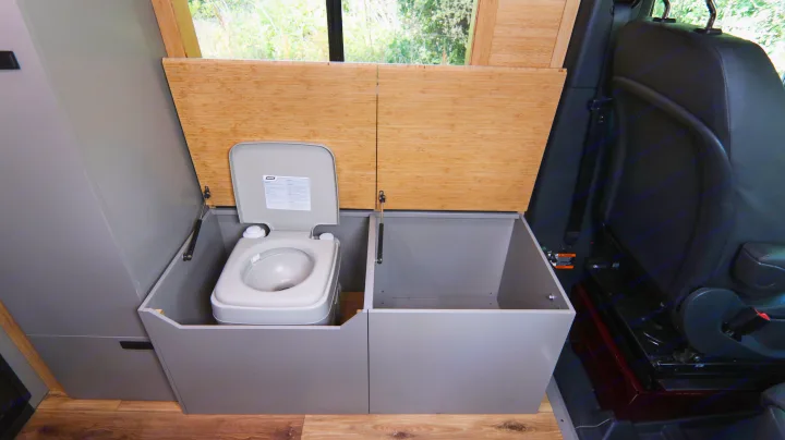 RV rental interior showcasing a compact portable toilet in a gray cabinet, with adjacent storage and bamboo lift-up lids.. Mercedes-Benz Sprinter 2025