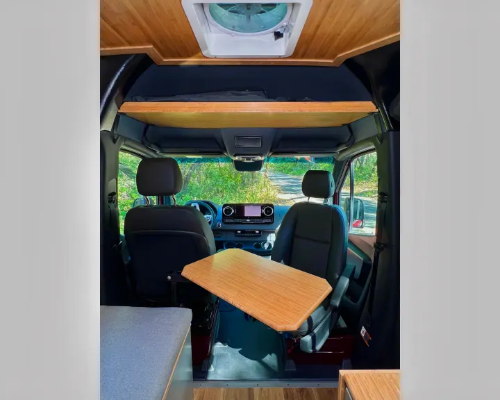 Inside this **rv rental** camper van: front seats, a versatile wooden table, and an overhead bunk, ready for your road trip.. Mercedes-Benz Sprinter 2025