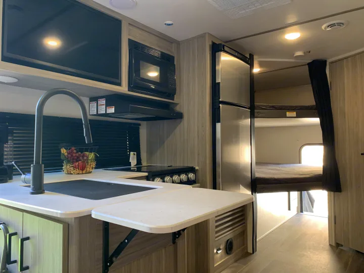 Modern rv rental interior: bright kitchen with sink, stove, microwave, TV, fridge, and bunk beds visible in the background.. Keystone RV Passport 2022
