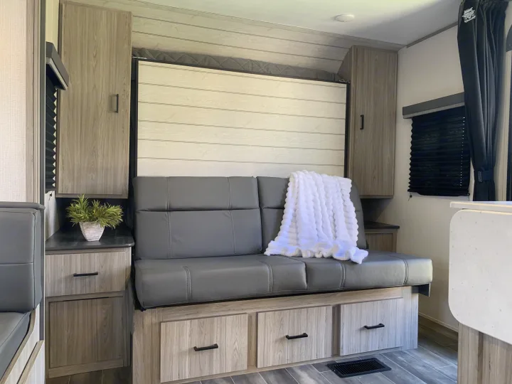 Cozy RV rental interior with a gray sofa, folded murphy bed, wooden storage, and a soft white blanket.. Keystone RV Passport 2022