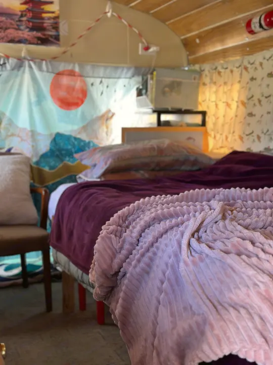 Inside a cozy RV rental: a plush bed with purple and pink blankets, nature tapestry, and wood-paneled ceiling.. GMC Vandura 1986