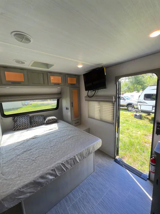 RV rental bedroom with a bed, patterned pillows, wall-mounted TV, and an open door to a sunny campsite.. Forest River Ibex 2021