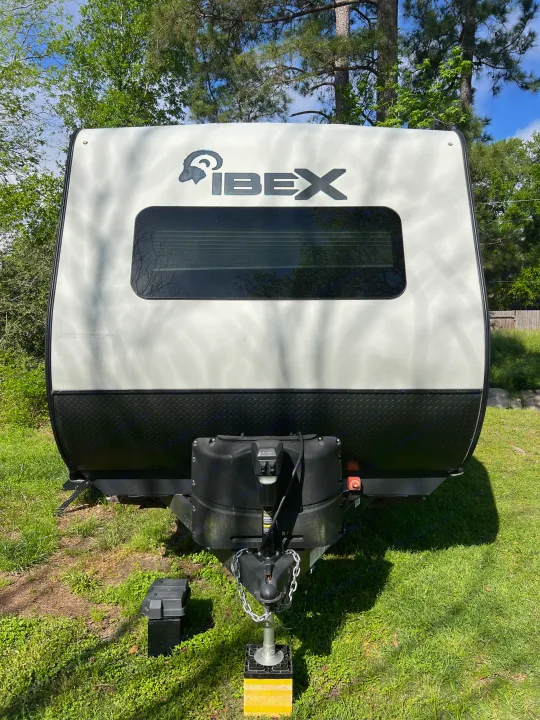 White and black Ibex travel trailer (RV rental) front view, showing its logo, front window, and hitch on grass.. Forest River Ibex 2021