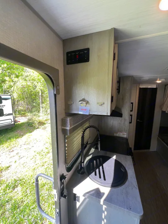 RV rental interior: Kitchen area with a sink, faucet, control panel, bottle opener. Open door reveals green outdoors.. Forest River Ibex 2021