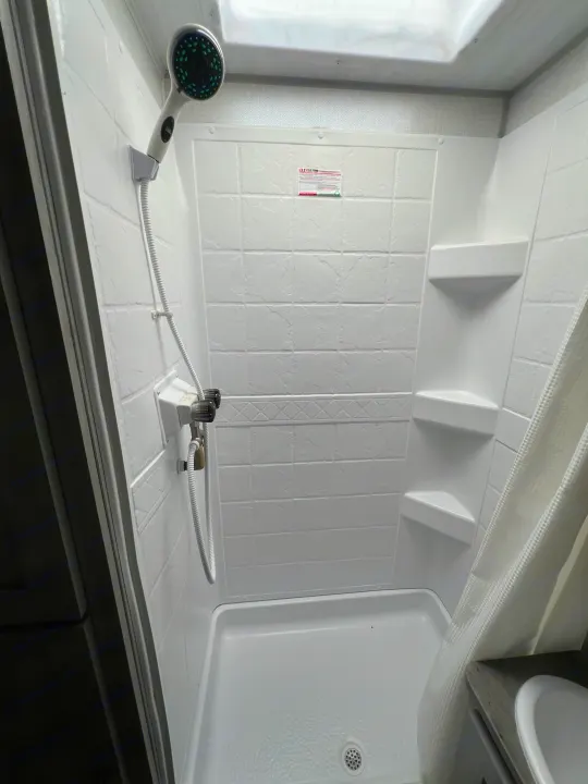 Inside an rv rental, a compact white shower stall with a handheld showerhead, two control knobs, corner shelves, and a drain.. Forest River Ibex 2021