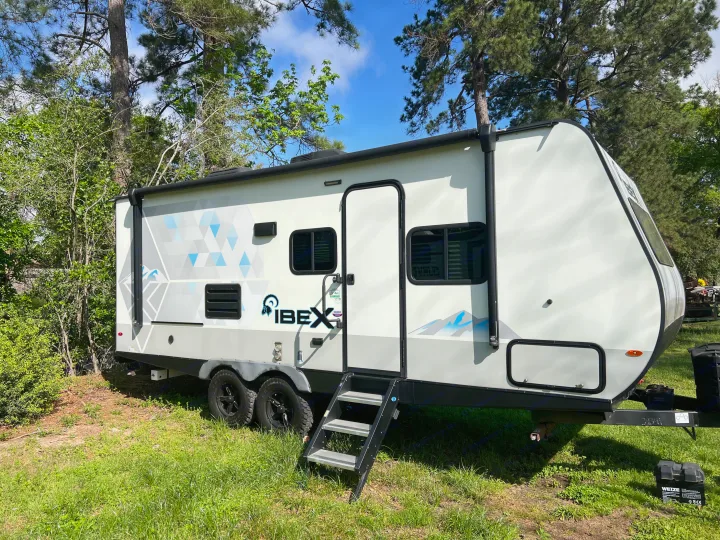 An Ibex rv rental travel trailer, white with blue/grey graphics, parked in a sunny, wooded campsite.. Forest River Ibex 2021