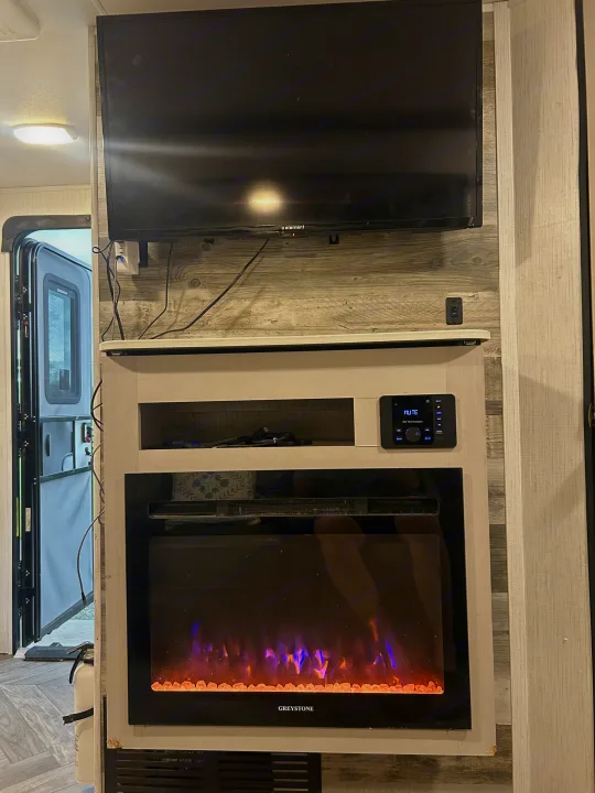 Inside an RV rental: a wall-mounted TV, electric fireplace (Greystone) with glowing flames, and an IRV media player.. Heartland RVs Mallard 2023