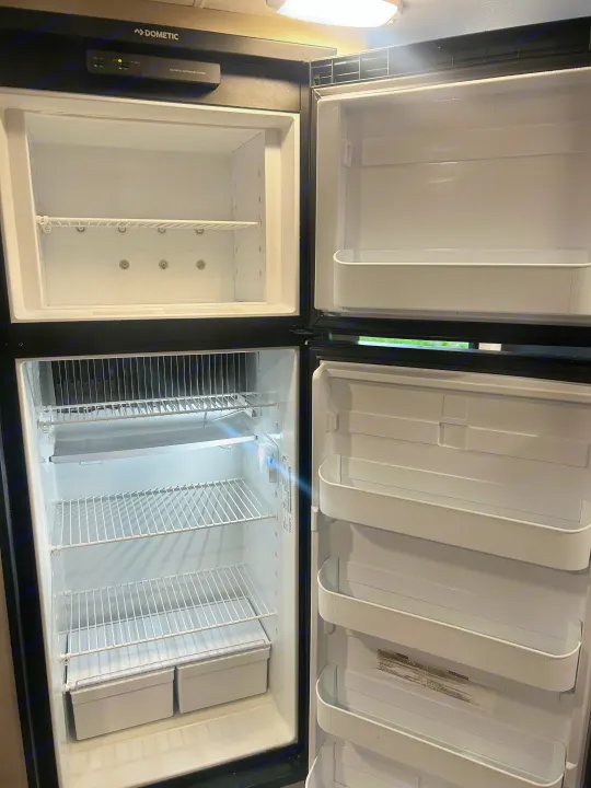 Dometic RV rental refrigerator open and empty, showing its freezer, fridge, shelves, and door storage.. Heartland RVs Mallard 2023