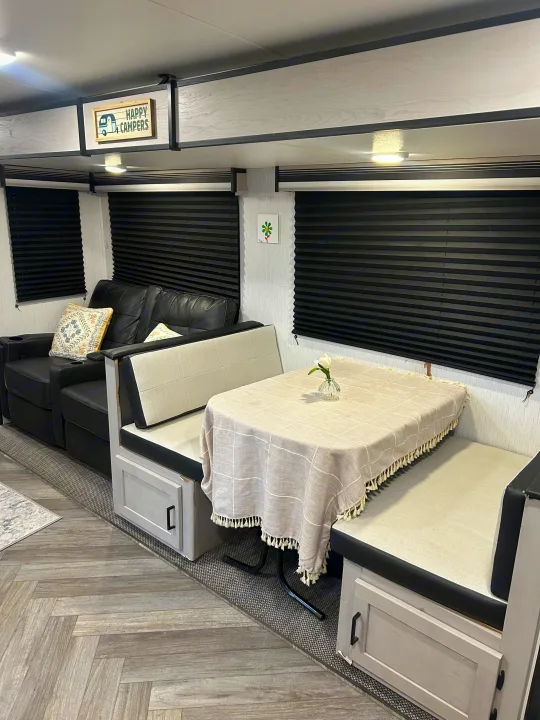Cozy rv rental interior featuring a black sofa, dining booth with tablecloth, and cheerful "Happy Campers" sign.. Heartland RVs Mallard 2023