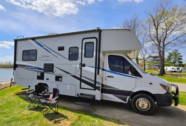 A Coachmen Prism RV rental is set up lakeside with chairs, offering a scenic view. Water park & other RVs in distance.. Forest River Coachmen Prism 2020