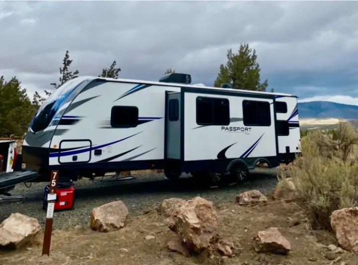 This RV rental, a white and blue travel trailer with an extended slide-out, is parked at a rugged campsite (site 75) under a cloudy sky.. Keystone RV Passport GT 2021