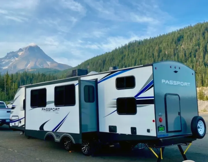 White & gray Passport RV rental, with an open slide-out, parked in front of a majestic mountain and dense forest.. Keystone RV Passport GT 2021