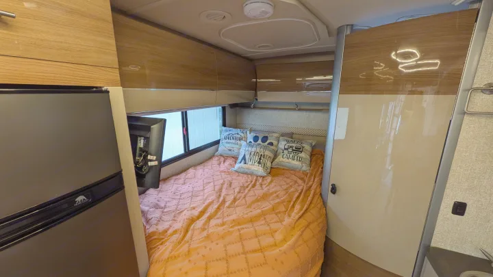 Cozy RV rental bedroom featuring an orange bed with decorative pillows, a window, overhead cabinets, and a refrigerator.. Winnebago View 2017