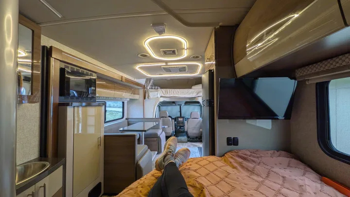 Cozy RV rental interior with feet up on the bed. Features a kitchen, dinette, TV, & bright lighting for travelers.. Winnebago View 2017