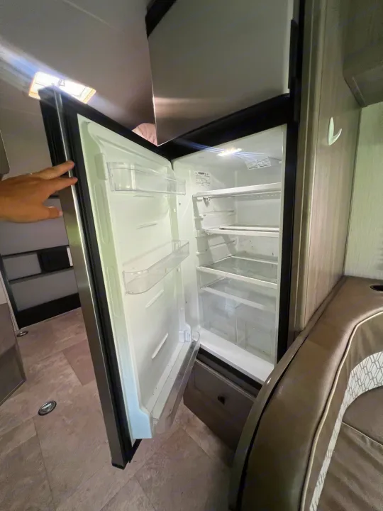 Compact refrigerator in an RV rental, shown open with a hand, revealing clear shelves and door storage inside.. Coachmen Freelander 22xg 2020