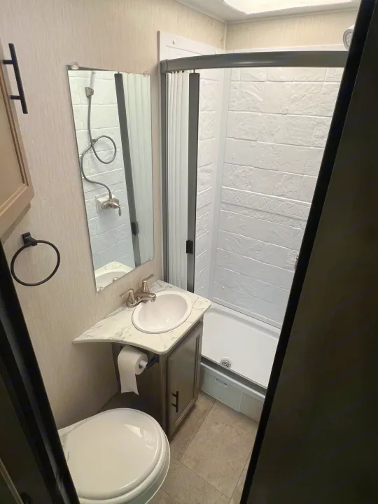 This RV rental bathroom features a white toilet, marble-look vanity with sink and mirror, plus an enclosed standing shower.. Coachmen Freelander 22xg 2020