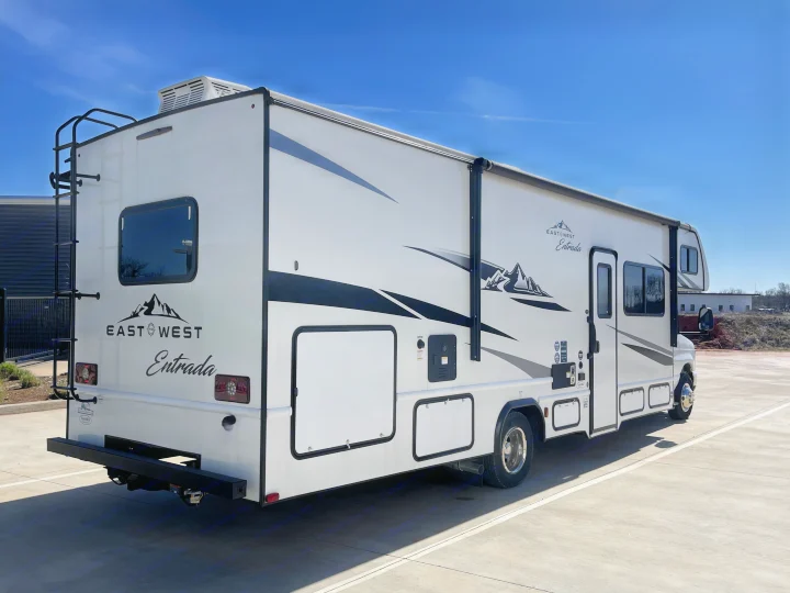 Side and rear view of a white East West Entrada Class C rv rental, featuring a rear ladder, side awning, and mountain graphics under a clear sky.. East to West Entrada 2024