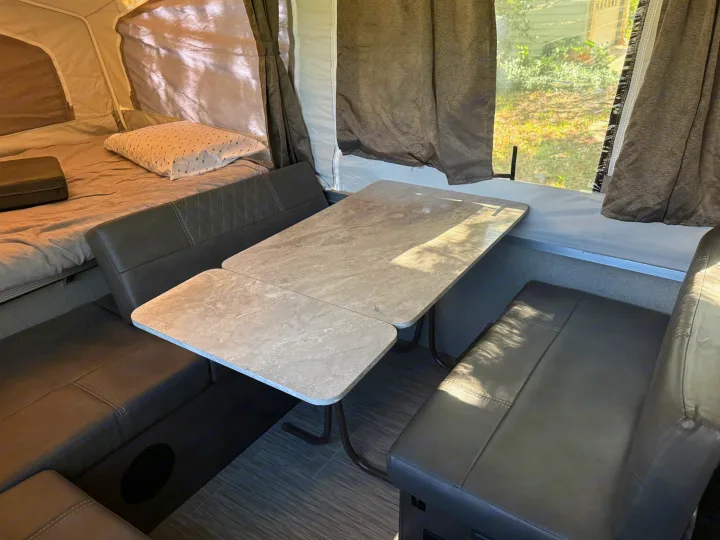 Inside this RV rental pop-up camper: a dinette area with a light patterned table and dark gray benches, next to a bed.. Forest River Flagstaff 2020