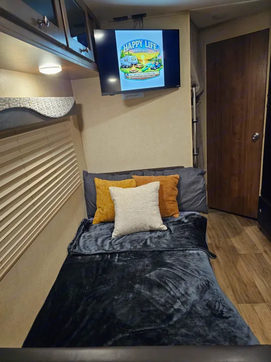 This cozy RV rental features a bedroom with a comfortable bed, overhead bunk, and a TV displaying the Happy Life Rentals logo.. Aspen Trail Aspen Trail Trailer 2018