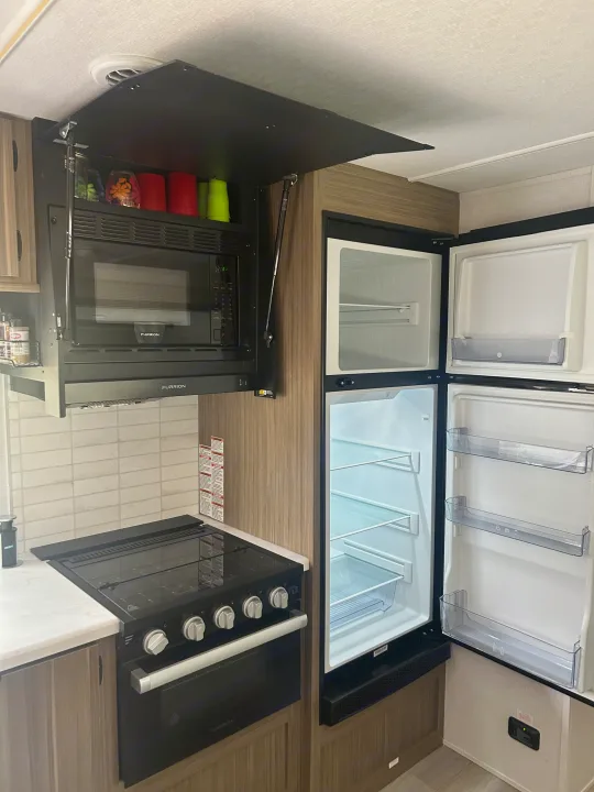 Modern RV rental kitchen featuring a Furrion microwave, stove, and a fully open, clean refrigerator with shelves, ready for use.. Forest River Wildwood 2025