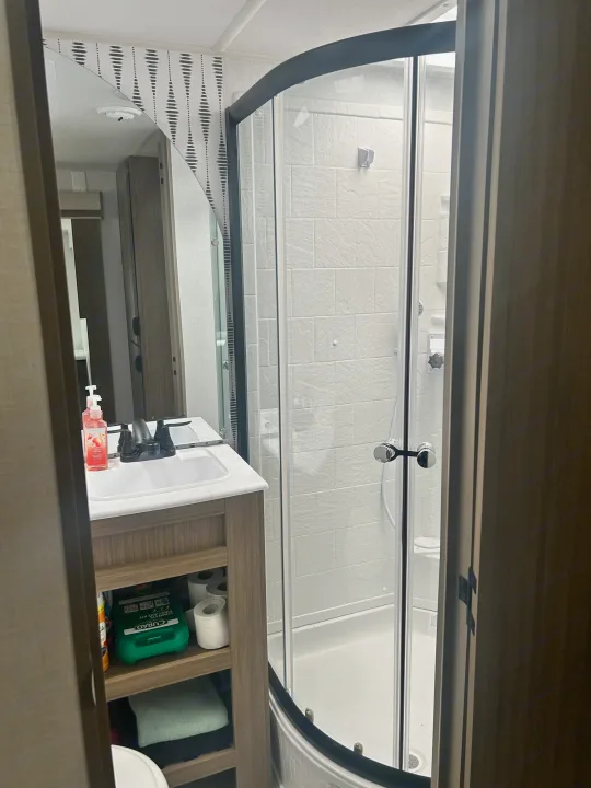RV rental bathroom: Sink, mirror, and curved shower with white tiles. Shelves hold essentials like first aid and toilet paper.. Forest River Wildwood 2025