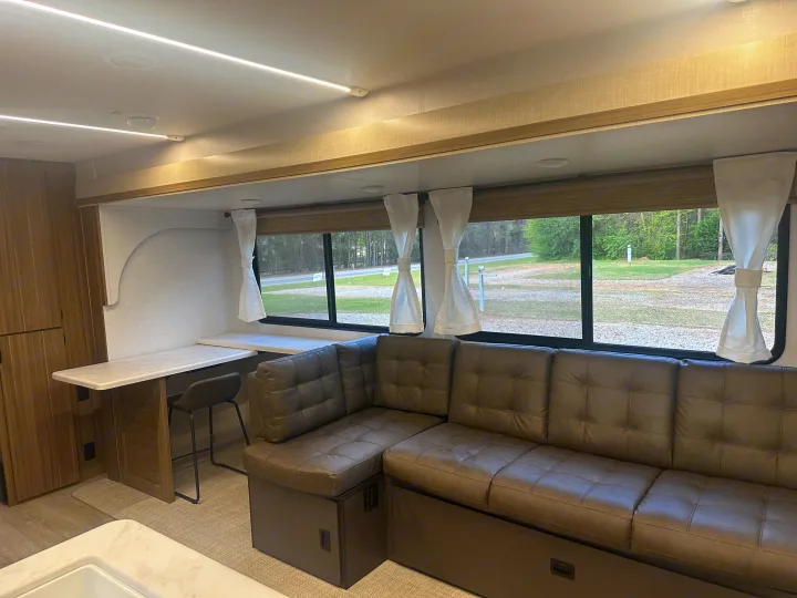 Spacious rv rental interior featuring a brown L-shaped couch, desk, and large window with an outdoor view.. Forest River Wildwood 2025