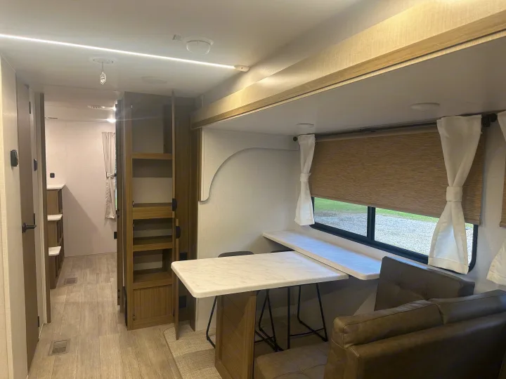 Modern RV rental interior, showing a dinette table & seating by a window, with a hallway & storage visible.. Forest River Wildwood 2025