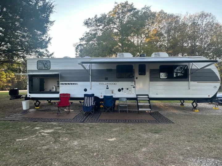 This RV rental is set up at a campsite, featuring an extended awning, outdoor kitchen, and chairs on a mat.. Forest River Wildwood 2025