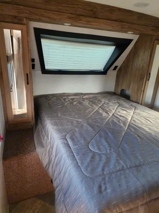 Step into this cozy RV rental bedroom featuring a grey bed, large window with blinds, and warm wood accents.. Palomino Other 2022