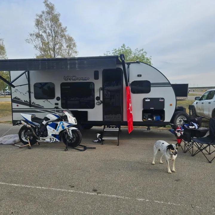 This RV rental setup shows a Wolf Pup travel trailer with an extended awning, a racing motorcycle, and a spotted dog at a track.. Forest River Cherokee Wolf Pup 2020