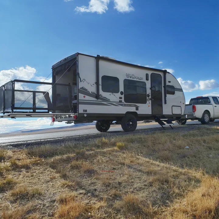 Sunny day with an RV rental: a Wolf Pup toy hauler with its rear patio deck open, hitched to a truck, on a road with dry grass.. Forest River Cherokee Wolf Pup 2020