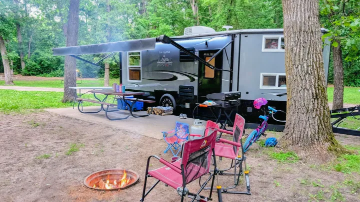 Relaxing RV rental campsite setup: gray camper, fire pit with burning wood, grill, picnic table, and chairs under trees.. Ice Castle Fish Houses Ice Castle Fish Houses Trailer 2023