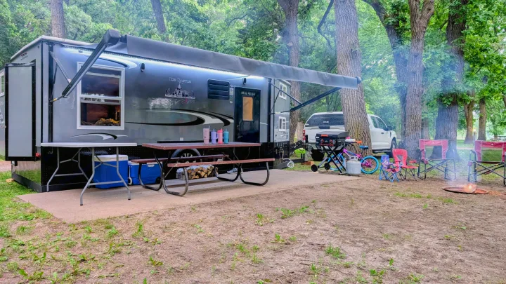 This spacious RV rental is set up at a wooded campsite with a campfire, picnic table, and chairs. Perfect outdoor adventure!. Ice Castle Fish Houses Ice Castle Fish Houses Trailer 2023