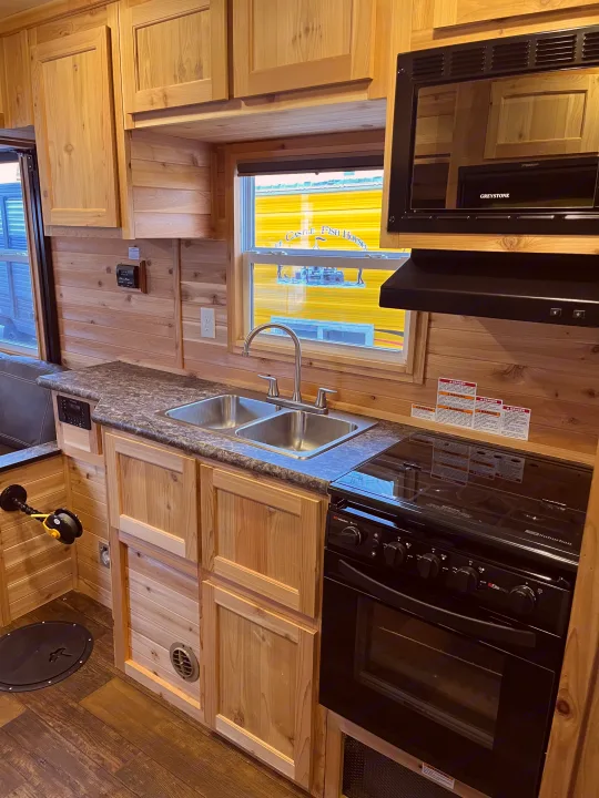 Cozy RV rental fish house kitchen featuring light wood paneling, a double sink, black stove, microwave, and a floor fishing hole.. Ice Castle Fish Houses Ice Castle Fish Houses Trailer 2023
