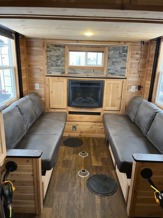 Cozy RV rental interior: wood-paneled ice fishing house with benches, fireplace, and fishing holes on the floor.. Ice Castle Fish Houses Ice Castle Fish Houses Trailer 2023