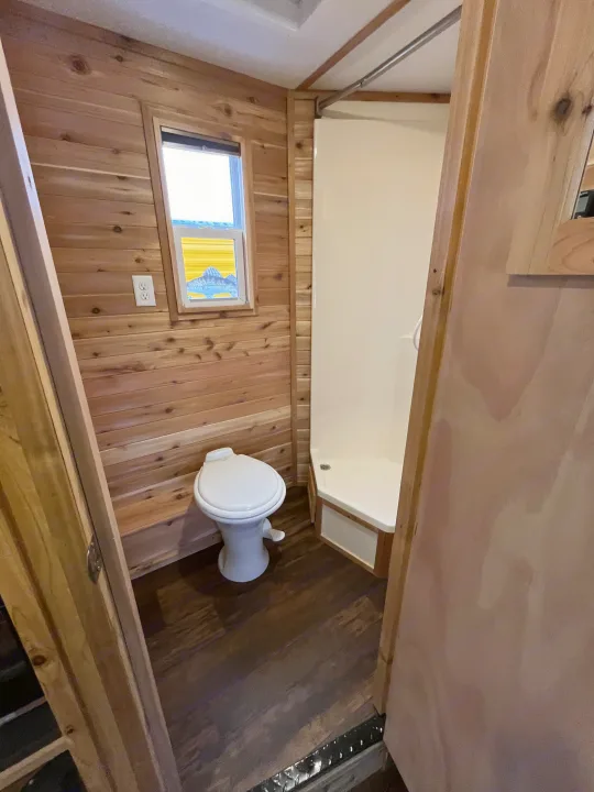RV rental's compact bathroom features rustic wood walls, a white toilet, and a corner shower unit, ideal for travel.. Ice Castle Fish Houses Ice Castle Fish Houses Trailer 2023