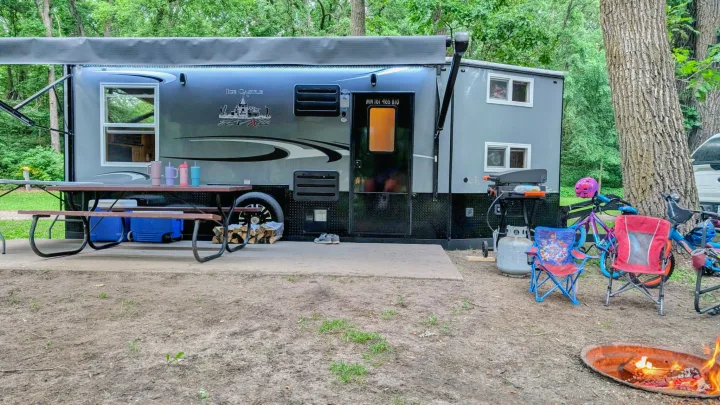 RV rental camper set up at a wooded campsite with a picnic table, grill, bikes, and a cozy fire pit.. Ice Castle Fish Houses Ice Castle Fish Houses Trailer 2023