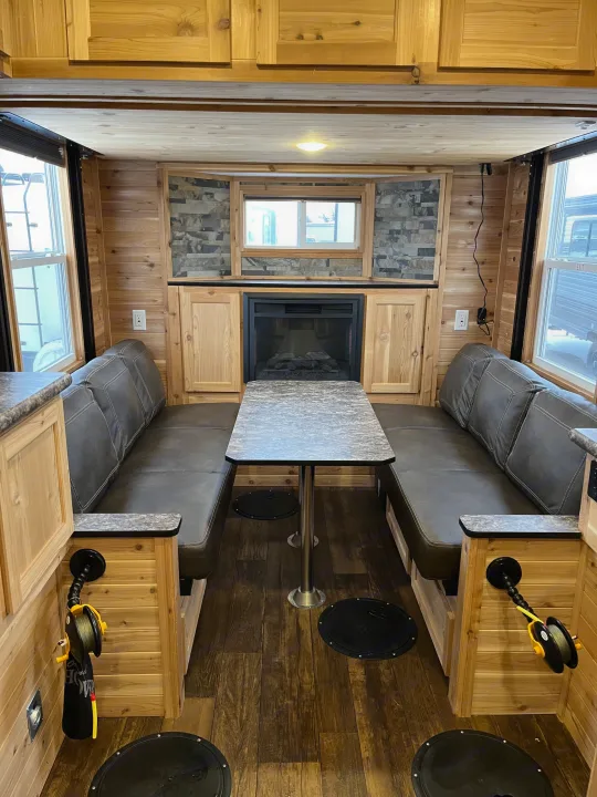 Spacious RV rental interior: wood-paneled walls, dining area with benches, fireplace, and covered ice fishing holes on the floor.. Ice Castle Fish Houses Ice Castle Fish Houses Trailer 2023