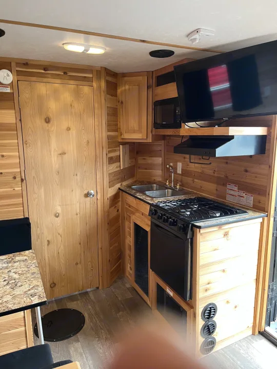Cozy RV rental interior with wooden walls. Features a kitchen area with double sink, stove, microwave, and a wall-mounted TV.. Ice Castle Fish Houses Ice Castle Fish Houses Trailer 2020