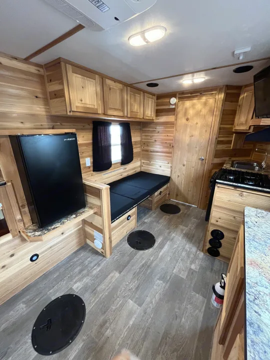 Cozy RV rental interior featuring a compact kitchen, bench bed, fridge, and practical floor hatches in a wood-paneled space.. Ice Castle Fish Houses Ice Castle Fish Houses Trailer 2020