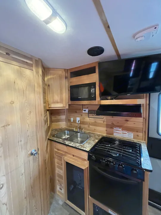 RV rental kitchen: light wood paneling, double sink, gas stove, microwave, and TV in a compact, well-equipped space.. Ice Castle Fish Houses Ice Castle Fish Houses Trailer 2020