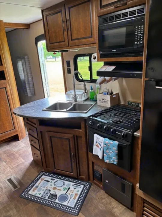 View inside an RV rental kitchen. Features include a double sink, microwave, stovetop, and storage cabinets. Green view outside.. Crossroads RV Zinger 2017